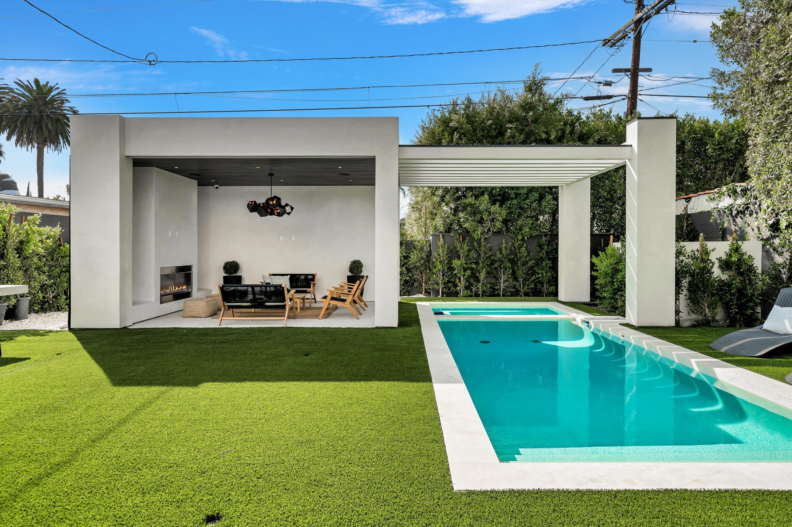 Modern backyard with a rectangular pool, artificial grass, and a covered seating area. The area is shaded by trees and the sky is clear.